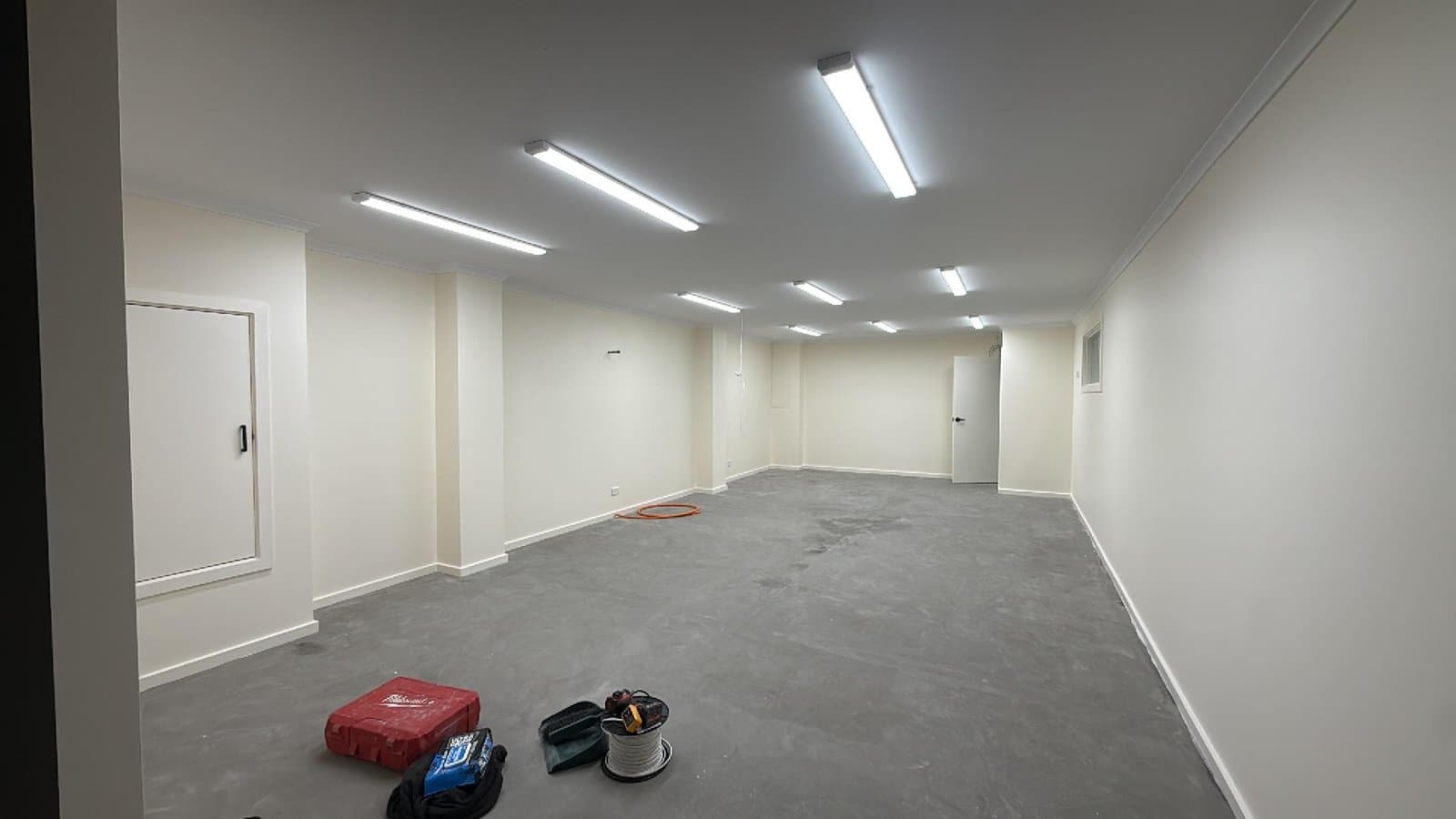Wide interior with rows of illuminated linear ceiling lights and work tools on the floor.